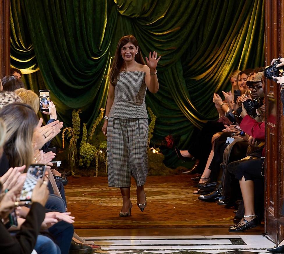 A woman in grey trousers and top, standing at the end of a runway in front of a large green curtain, waving at the crowd