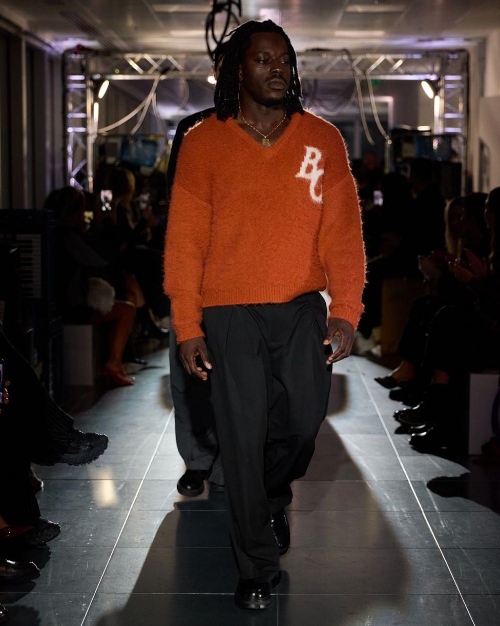 A man on a runway, wearing an orange jumper with white BG letters, and dark trousers.