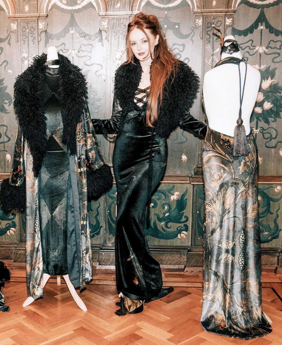 A woman with long ginger hair in a dramatic black dress and fur stole, she is stood beside two mannequins that have on similar outfits, inside a grand home.