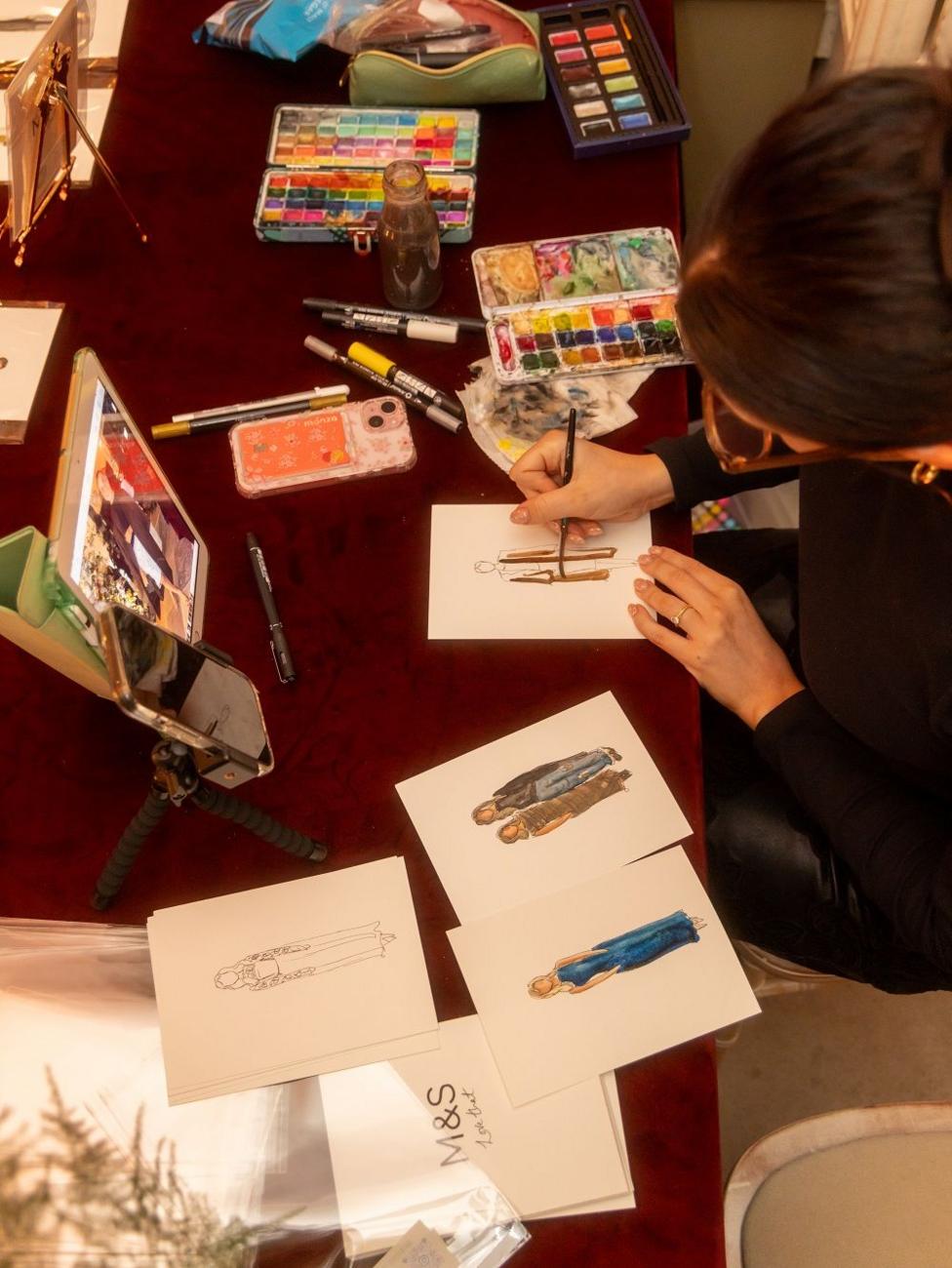 Aerial shot of someone sketching fashion illustrations on a busy table, with lots of paints and sketches on display