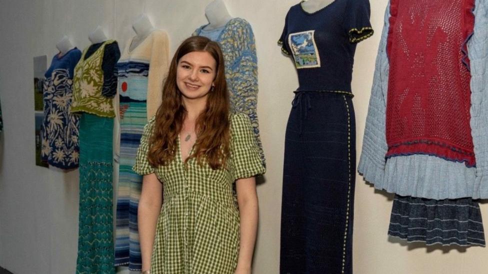 A girl with long brown hair, smiling athe the camera, shes wearing a green and yellow gingham checked dress. Stood in front of a range of hanging designs.