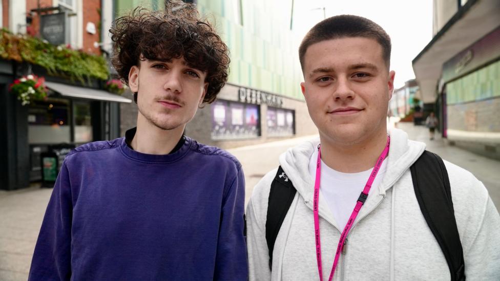 Two teenagers stand in a city centre street, both have dark hair and are looking at the camera.