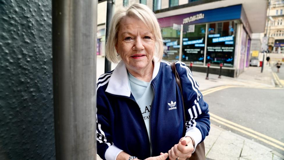 A woman in a navy jacket with white stripes on the arms stands in a city centre street, she has blonde her to her chin.