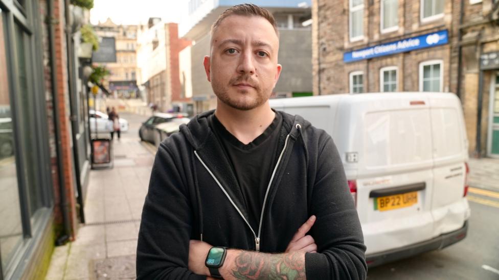 A man in a black zip-up jacket with tattoos visible on his forearm stands in a city centre street looking at the camera, he has dark blonde hair.