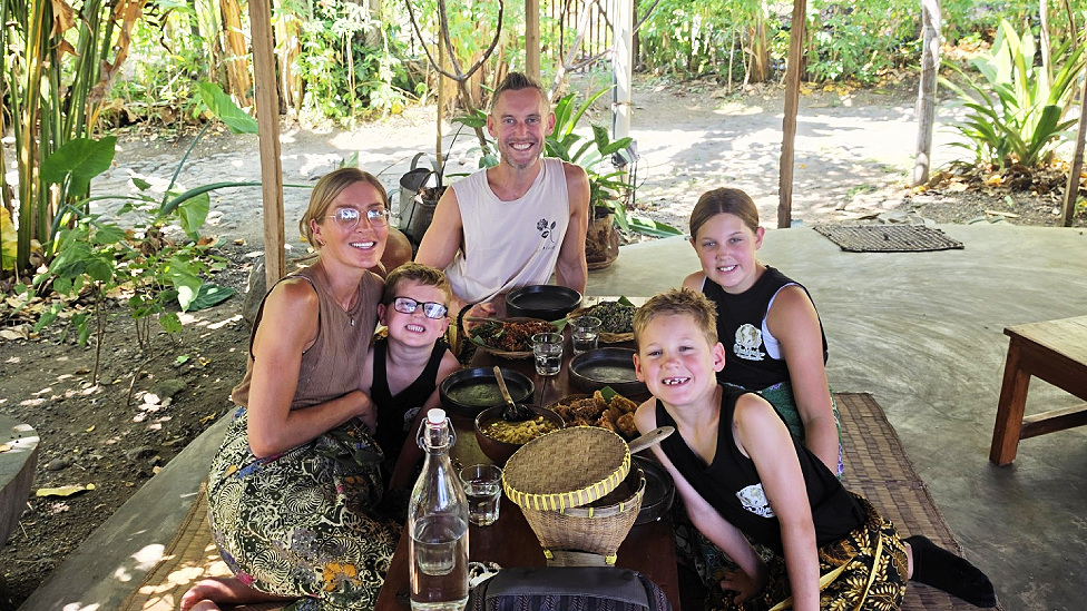 Five Woolridges sitting under a woven canopy on a mat on the floor with woven containers of food and bowls. They are wearing traditional material as skirt-type wraps. All five are looking at the camera and smiling.