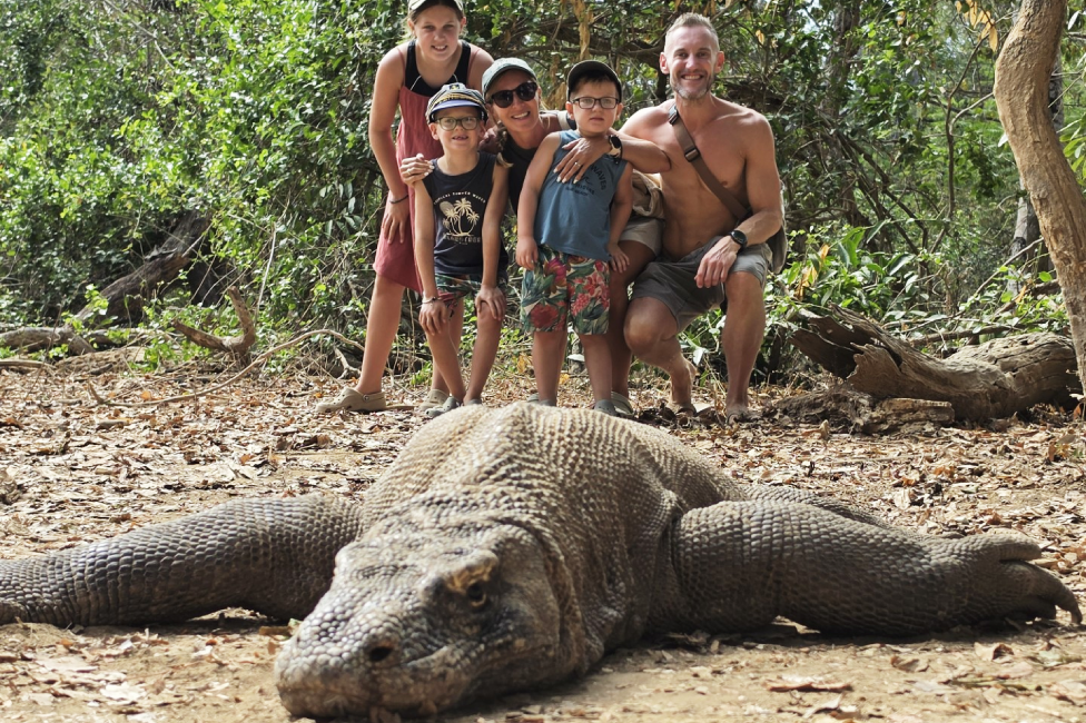 A large Komodo dragon is flat on the ground in the foreground of the photo with the family of five pictured behind it. There are trees and logs in the background.