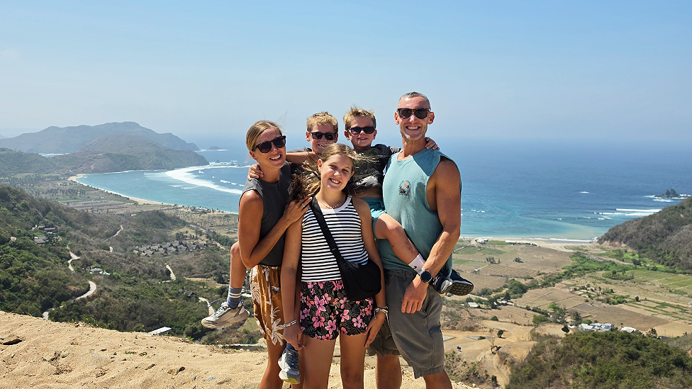 Five family members huddled in a group. The two boys are help up by Dani and Tom on either side, with Olivia standing in front. All except Olivia wear sunglasses and they're all wearing shorts and sleeveless t-shirts. They stand on a sandy hill up above fields with a narrow strip of beach and sea behind them. It is sunny and slightly hazy.