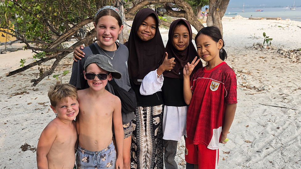 George, Morgan and Olivia stand next to three local Indonesian girls on a beach. Two of the girls wear black headscarfs and local clothing. The third wears a red and white football strip. The boys wear long beach shorts and Olivia wears a t-shirt and short and a bandana in her hair. They are standing underneath a tree.