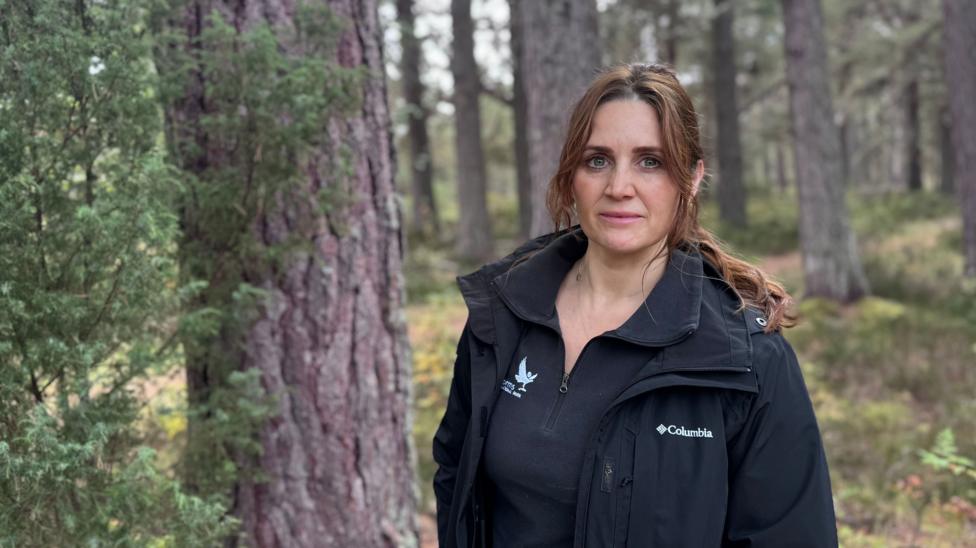 A woman standing in woodland, facing the camera. She has long brown hair tied back in a ponytail, wearing a black waterproof jacket and a black fleece.