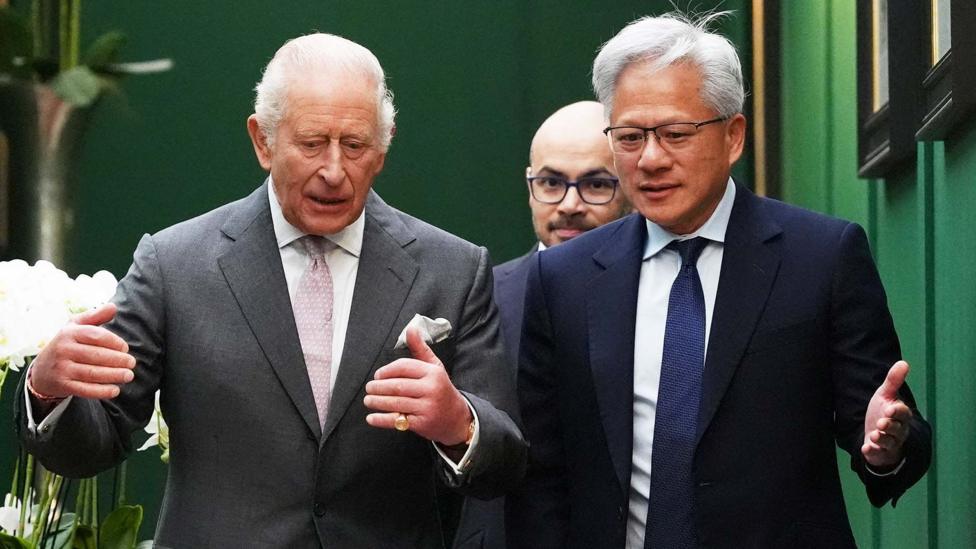 King Charles wearing a grey suit jacket and white shirt and pink tie, talking to Jensen Huang, the CEO of Nvidia, who is wearing a dark suit, white shirt and blue tie. Former Google DeepMind boss Sir Demis Hassabis is walking behind them.