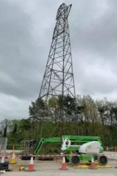 A tall electricity pylon, surrounded by building site equipment, tape and cones. Trees are visible behind it.