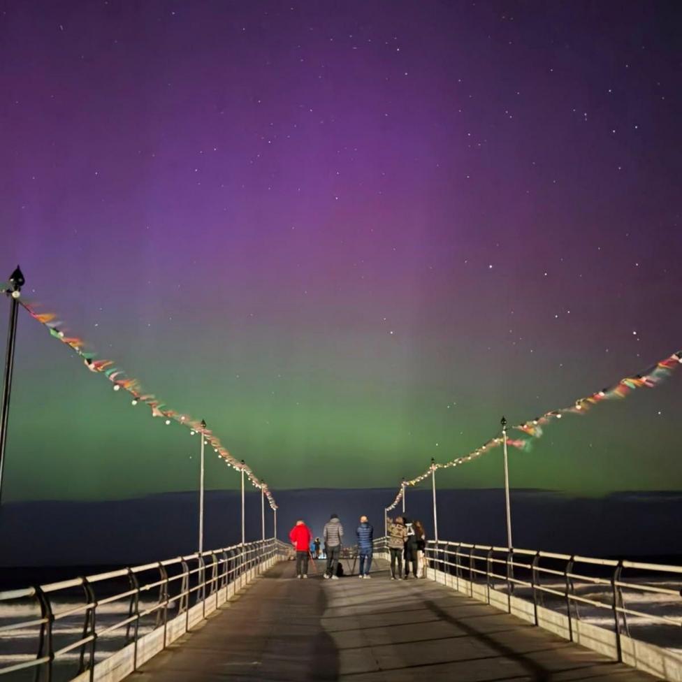 An image of the sky in Saltburn-by-the-Sea, Redcar and Cleveland, on 10 October during the evening. The Northern Lights can be seen in a pink and green hue alongside five people standing on a walkway at the bottom of the image.
