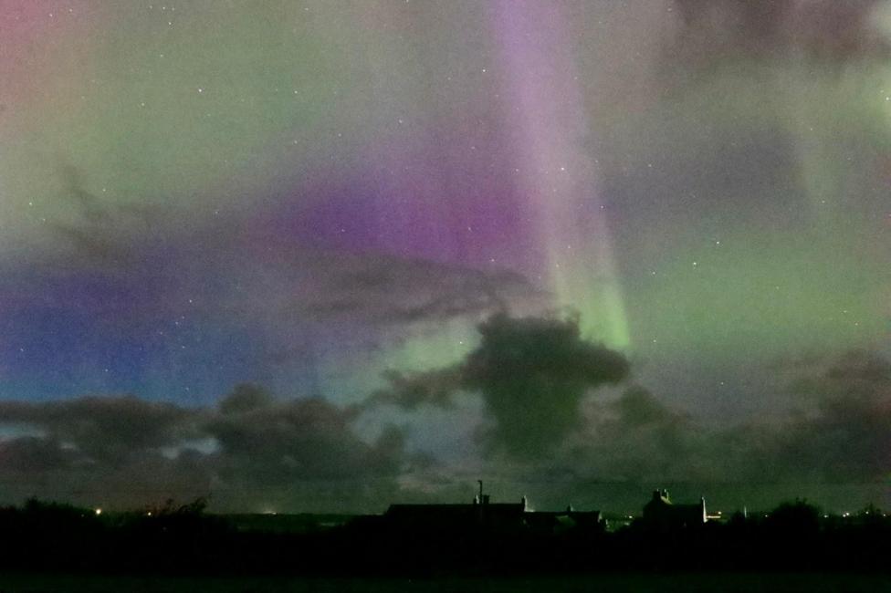 An image of the sky in Stenness, Orkney Islands, on 10 October during the evening. The Northern Lights can be seen in a pink and green hue with clouds visible above a skyline of houses.