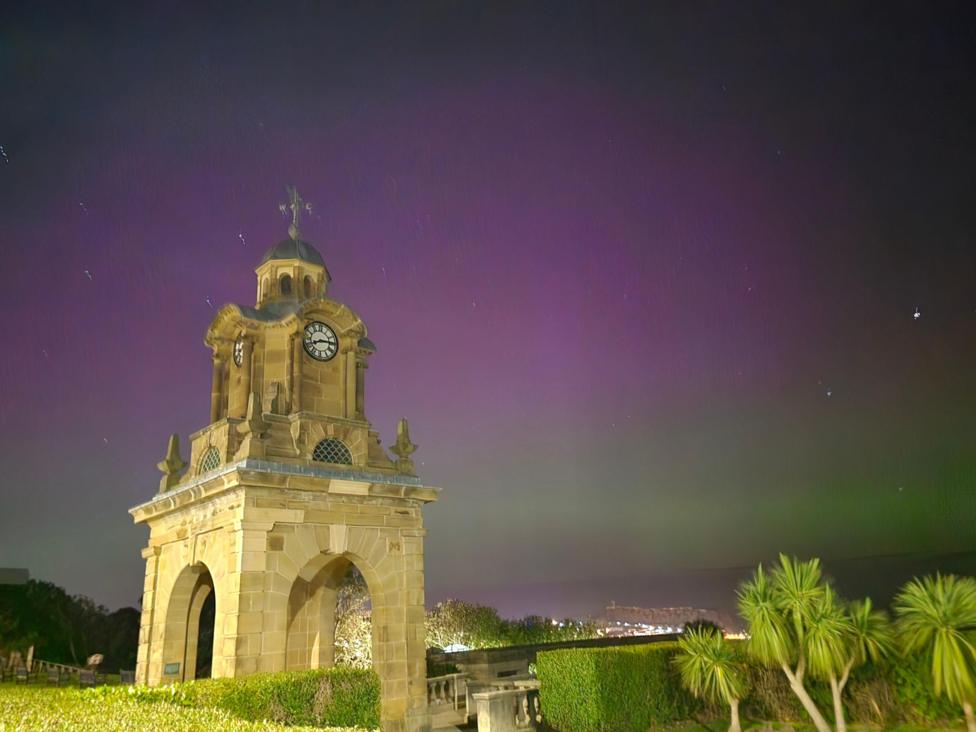 An image of the sky in Scarborough, North Yorkshire, on 10 October during the evening. The Northern Lights can be seen in a pink hue above a bell tower and green plants below.