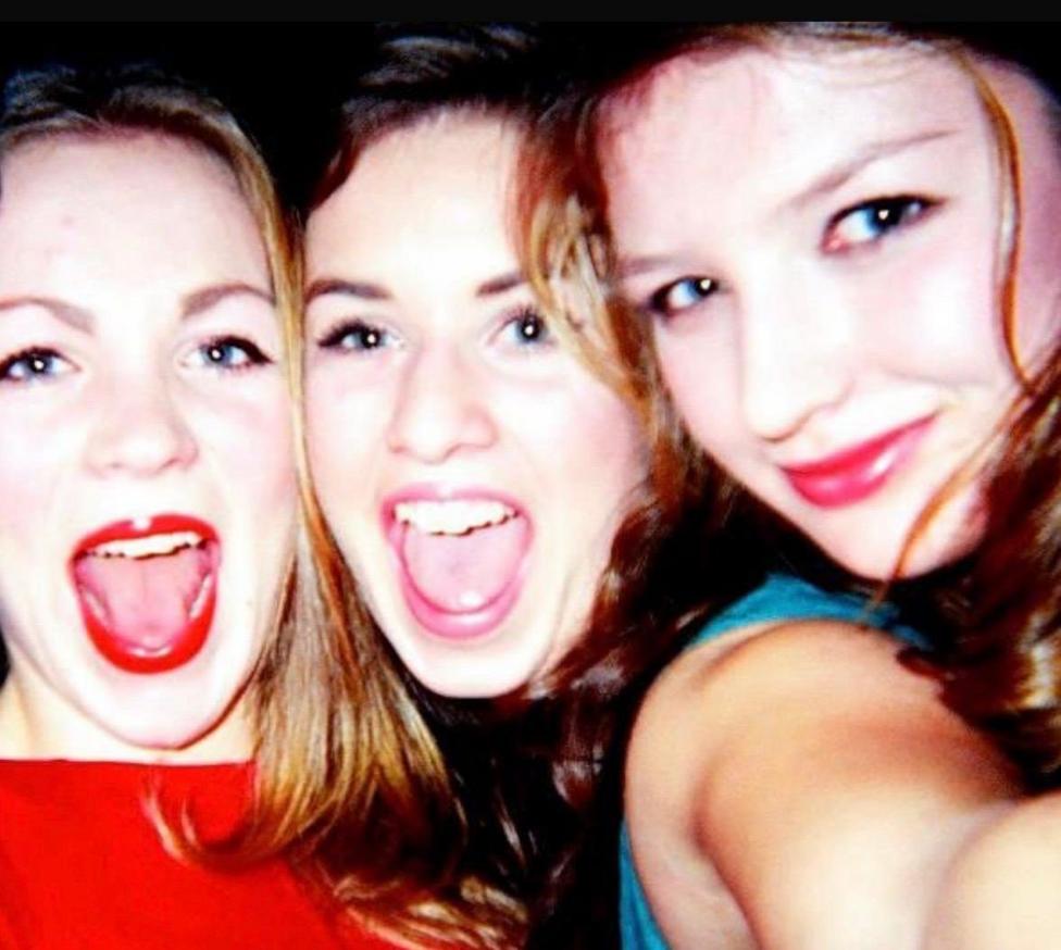 Three young woman pose close together. Lizzy and Charlotte (left and centre) have their mouths wide open, with Charlotte stretching out her arm to take the selfie. Libby has her mouth closed in a smile. They all have long brown hair