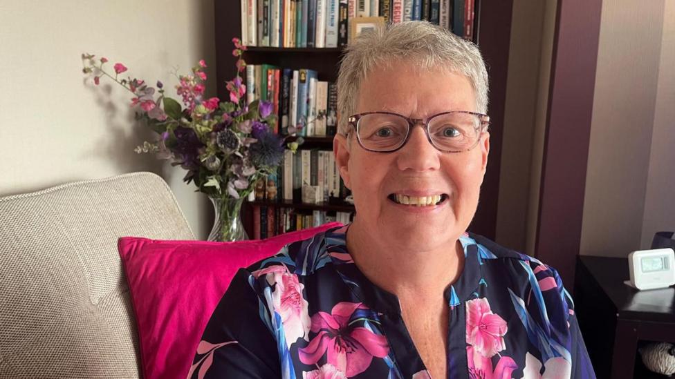 Woman wearing glasses and a floral-patterned top seated indoors beside a bright pink cushion, with a vase of flowers and a bookcase in the background.