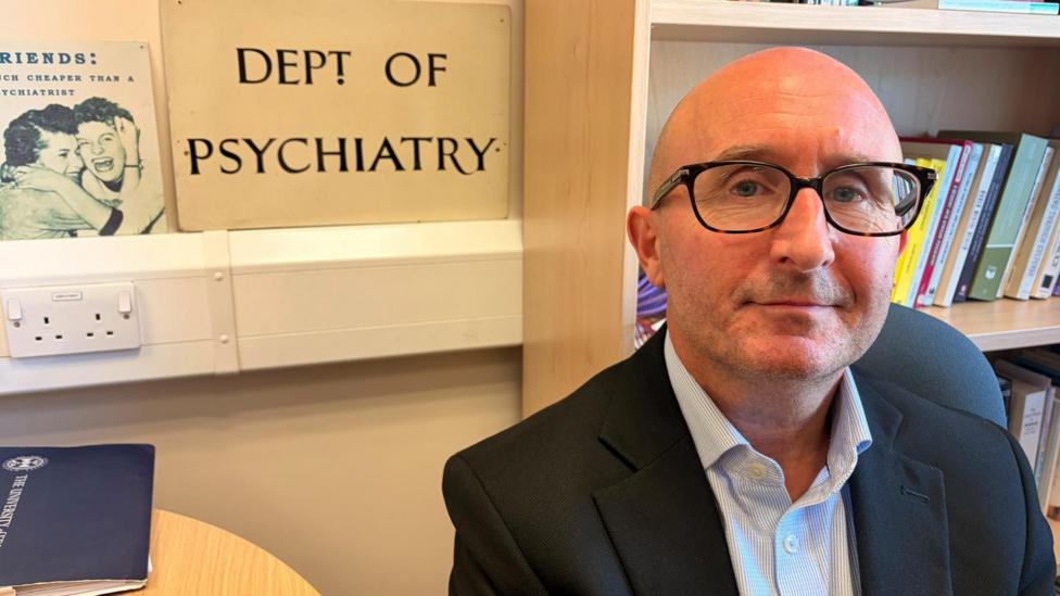 A man wearing glasses and a dark jacket seated in an office, with bookshelves and a sign reading “Dept of Psychiatry” visible in the background.