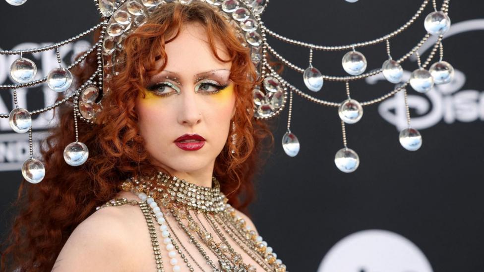 Close-up of a Chappell Roan wearing an ornate crystal headpiece and layered jeweled accessories, posing in front of a dark backdrop decorated with hanging reflective ornaments.