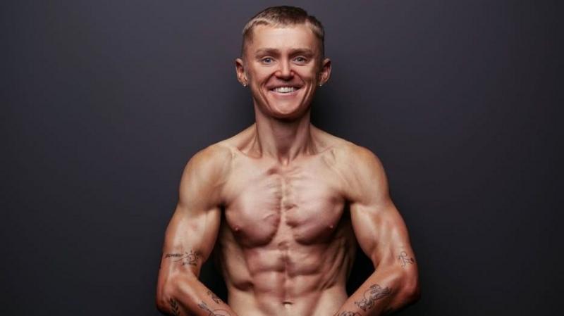 A man smiles at the camera, he has his top off posing