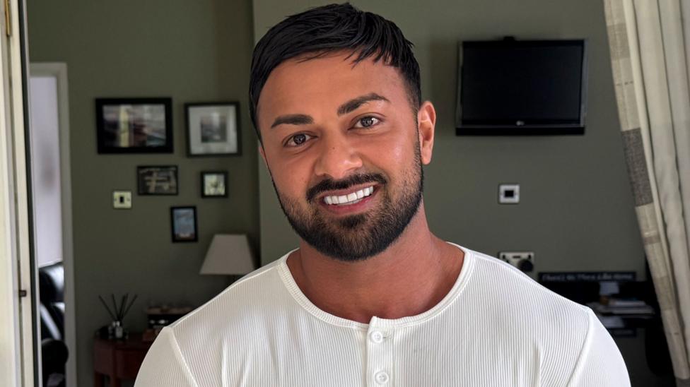 A man with dark hair and beard smiling at the camera, he is wearing a white top