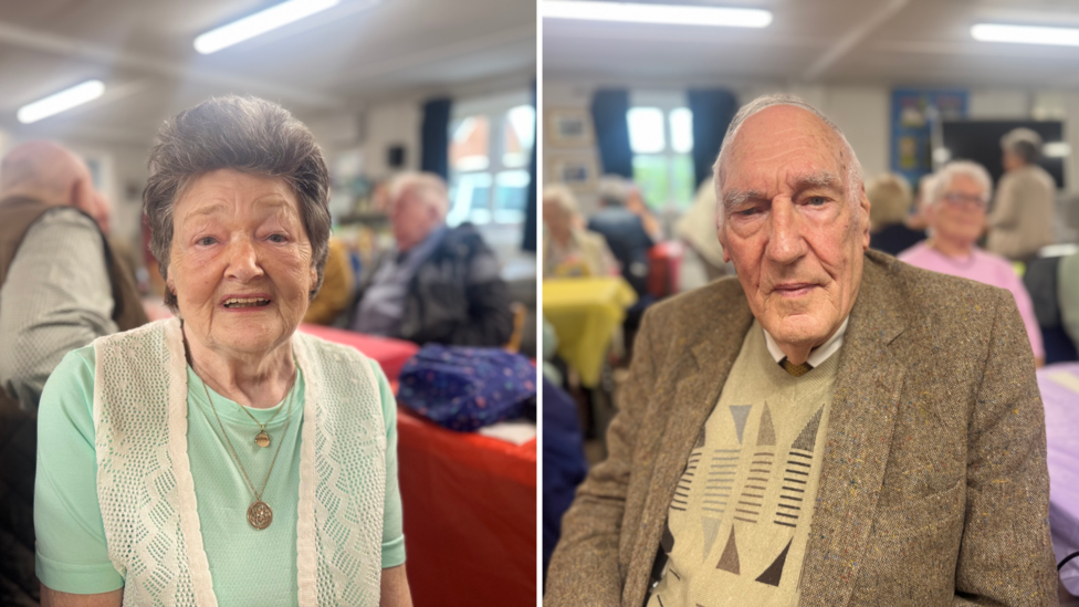 An elderly woman and an elderly man, pictured side by side. The woman has short dark grey hair and is wearing a light green t-shirt and white vest. The man has short grey hair and is wearing a beige jumper and a beige suit jacket.