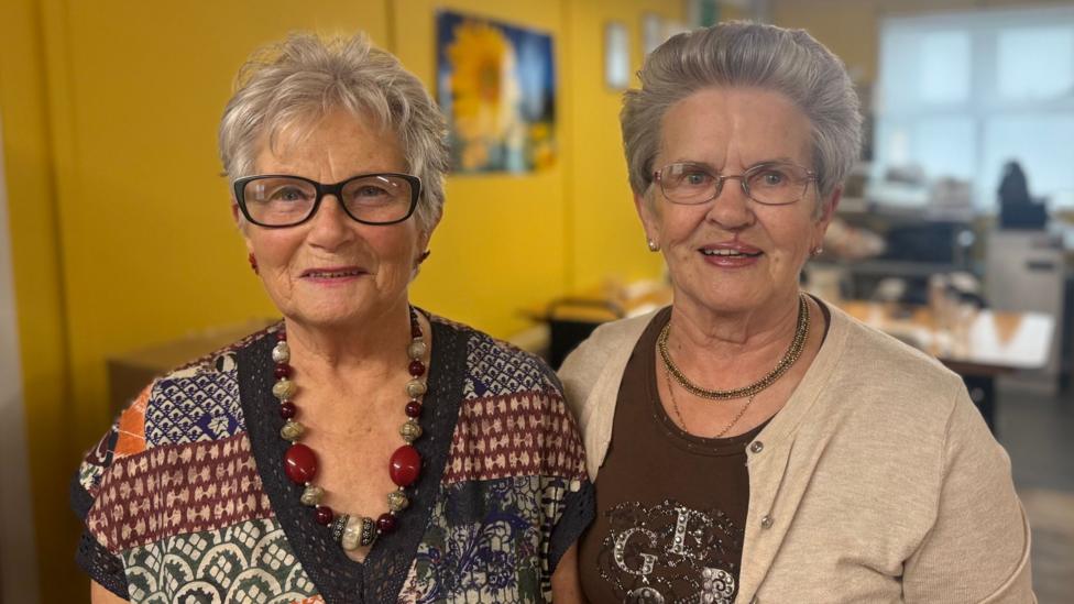 Two older women, standing side by side. They both have short grey hair and are both wearing glasses.