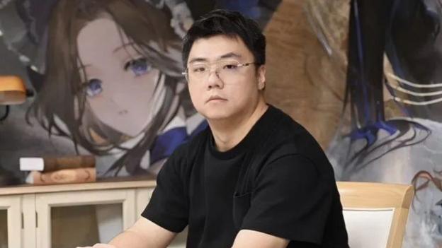 A man wearing glasses sat in a chair with a character from the game Reverse: 1999 on the wall behind.