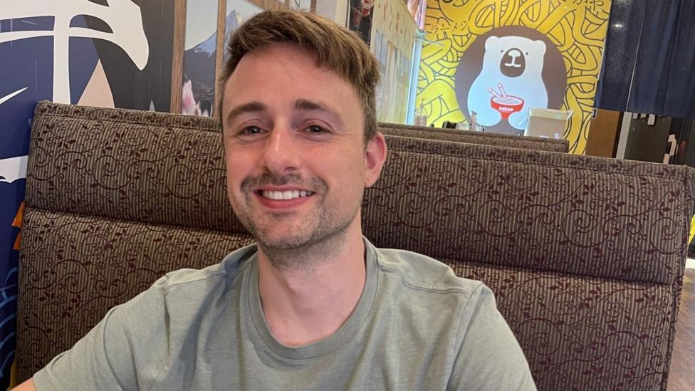 A guy with brown hair smiling sat on a sofa.