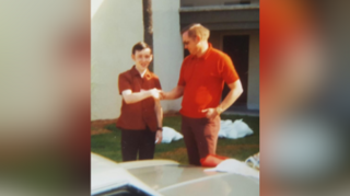 Gethings wears a red shirt and trousers and he stands shaking hands with Neil Armstrong, also in a red shirt and trousers - looking down at Gethins.