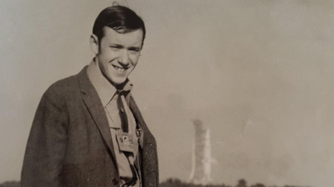 Gethings wears a shirt and jacket in front of a space launch pad. He smiles.