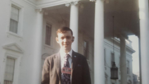 A picture of Gethins with short brown hair wearing a brown suit with a tie, standing in front of the White House.