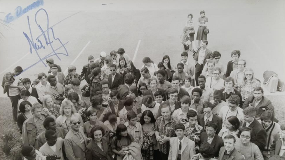 A black-and-white photograph of a large group of people shot at an aerial angle from above. It is signed 'To Dermot' accompanied by the autograph of Neil Armstrong.