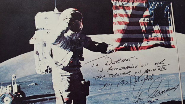 A postcard of an astronaut on the moon next to an American flag. The text on the card reads: 'To Dermot, In remembrance on our trip "together on Apollo XVII" My fondest wishes, Gene Cernan