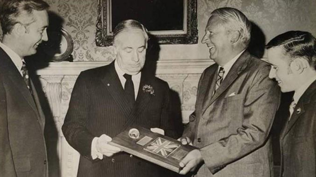 Three men stand smiling looking at a plaque with the union jack flag on it and a piece of rock. A teenage boy stands next to them.