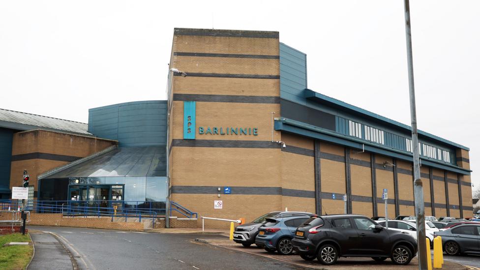 A large prison building, with a car park next to it. The building is sandstone coloured, with a sign saying Barlinnie in large letters on the wall