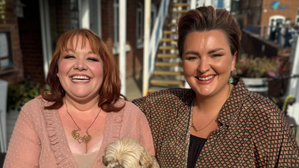 Jada Star (left) and Bronwen Lewis (right) are photographed outside sitting next to one another. Between them is a small white dog whose face is not in the photo. Star has read hair down to her shoulders. Lewis has short brown and red hair. They are both smiling and looking at the camera.