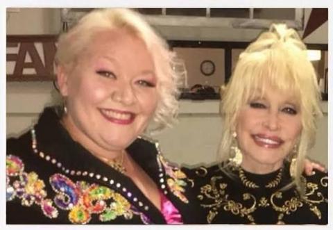 Jada Star and Dolly Parton are photographed standing next to one another. They both have blonde hair which is tied up. They are smiling and looking at the camera.