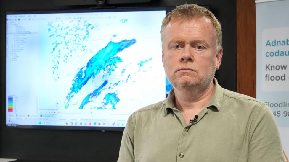 Jeremy Parr of Natural Resources Wales stands in front of a large screen displaying rainfall over a map of Wales. He has short, light brown hair and is wearing a light green shirt.