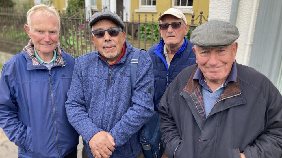 Friends Brian Churches, Jim Taylor, Dave Blake and Malcolm Butcher on the high street in Monmouth. They are all in their eightees and wearing a range of dark blue jackets. Jim Dave and Malcolm are wearing flat caps.