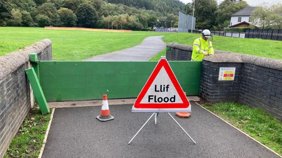 A green flood gate is closed by an NRW official wearing a bright yellow high-vis jacket and hard hat. There is parkland with trees in the background and a triangular warning sign in the foreground with the 'flood' on it.