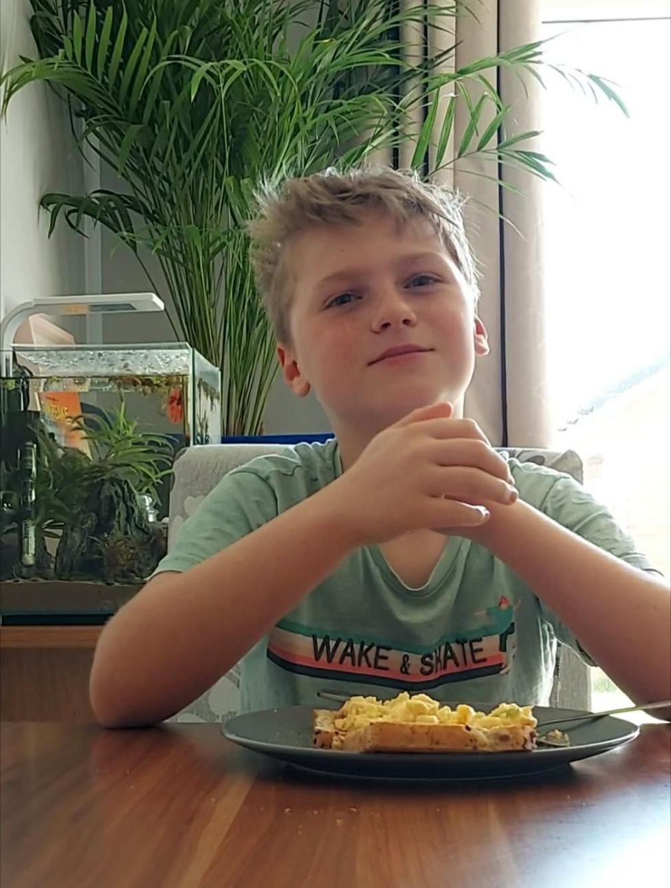 A little boy of around 11 years old sits at a table wearing a surf t-shirt clasping his hands above a plate of scrambled eggs on toast. There is a fish tank and a plant behind him to the left of the photo.