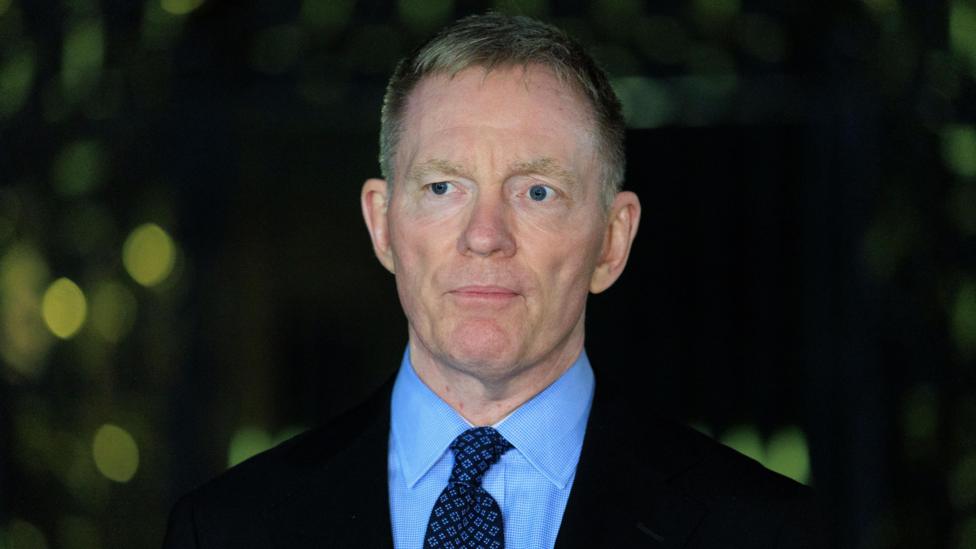 Sir Chris Bryant - man with grey hair, black blazer, blue shirt, and blue patterned tie. Backdrop behind him is dark.
