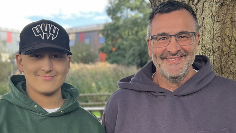 Ethan on the left and Lloyd on the right look at the camera and smile. Both are hoodies standing side by side outdoors beside a large tree. Ethan is wearing a black baseball cap with a white logo and Lloyd r wearing glasses. Trees, reeds and a building are out of focus in the background.