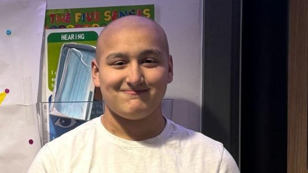 Ethan is standing with a toothless grin for the camera. He has on a white t-shirt and has close cropped hair. It is a head shot of him.
