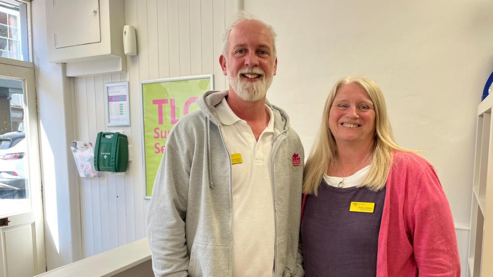 Two middle-aged people are pictured smiling in some kind of shop/office setting. The man is balding and has a white beard. He is wearing a white polo shirt under a light grey hoodie. The woman has long blonde hair and is wearing a dark purple tunic with a white T-shirt underneath and a bright pink cardigan over.