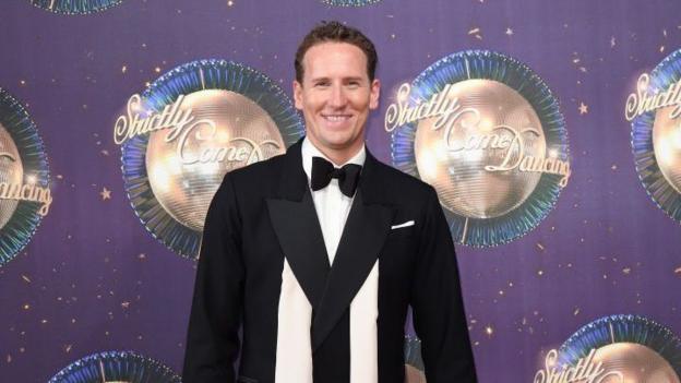 Brendan Cole attends the 'Strictly Come Dancing 2017' red carpet launch at Broadcasting House on August 28, 2017. He wears a black suit and black bow. He has brown hair and stands in front of a Strictly branded purple glitter ball background.