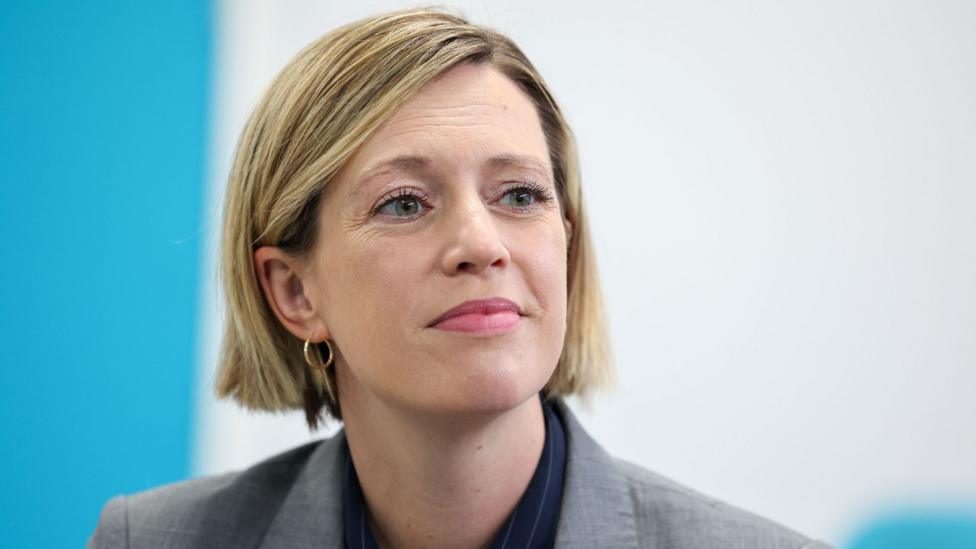 Jenny Gilruth has a blonde bob and is looking off to one side. She wears a dark shirt and grey blazer.