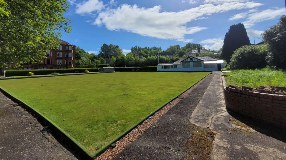A bowling green in the foreground with the white clubhouse in behind it. In the background are some flats to the left and trees.