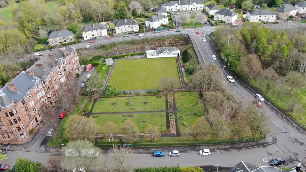 An aerial view of the site. The gardens are closest with the bowling green and clubhouse behind. Behind that is the train line and streets and houses. The site is bordered by more streets.