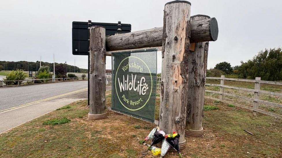 The park sign with some flowers laid by it. The sign is green fabric hung on a large wooden frame. There is grass all around and bunches of flowers at the scene.